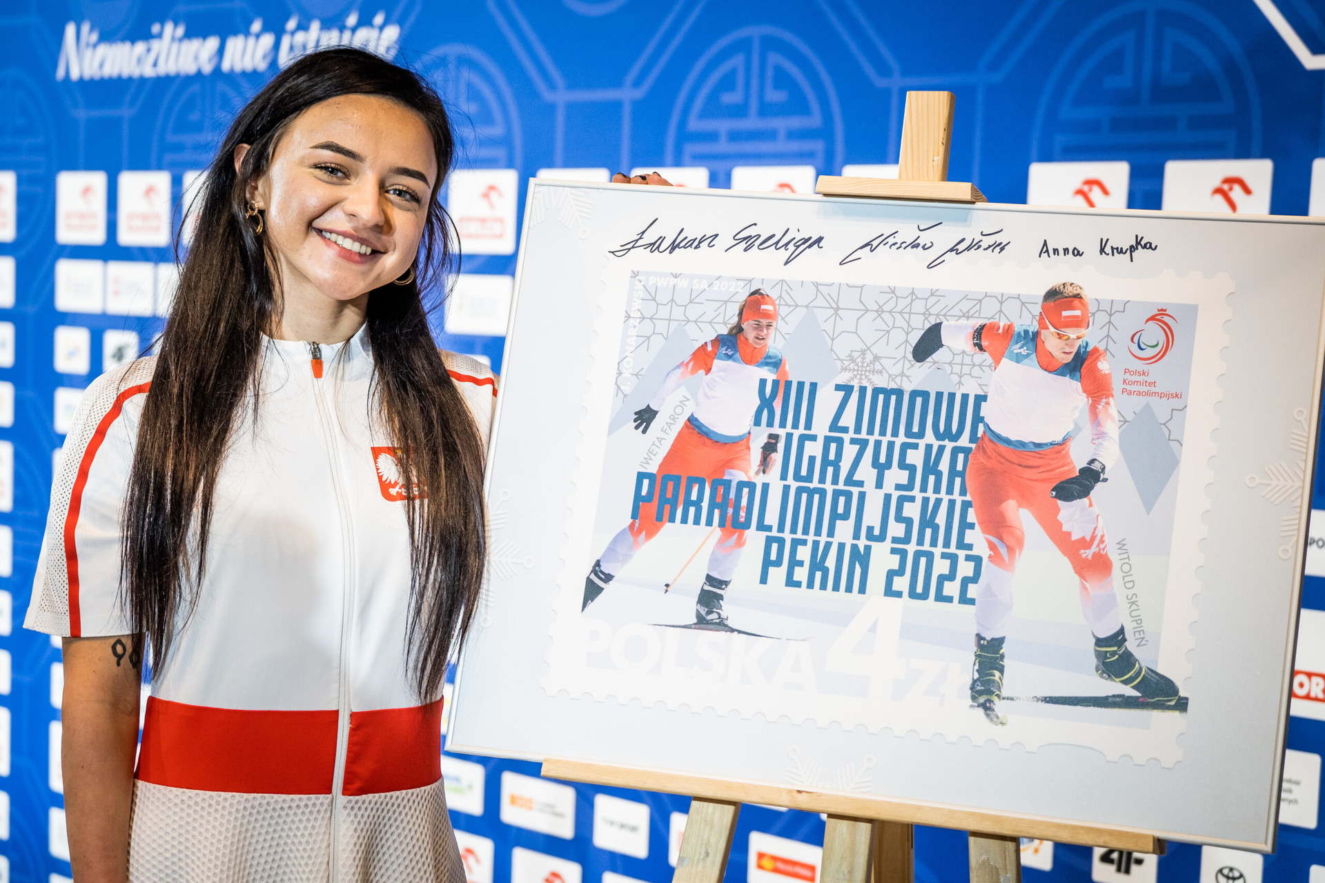 Zdjęcie przedstawia kobietę przy tablicy, na której jest logo XIII Zimowych Igrzysk Paraolimpijskich Pekin 2022