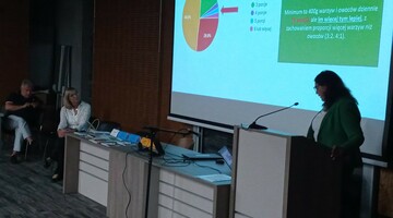 Pani Profesor prowadzi prezentację na konferencji, stojąc przy stole z laptopem i patrząc na publiczność.