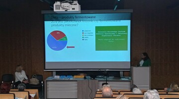 Sala wykładowa z osobami siedzącymi na krzesłach i ekranem projektora na przodzie.