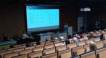 Sala wykładowa z osobami siedzącymi na krzesłach i ekranem projektora na przodzie.